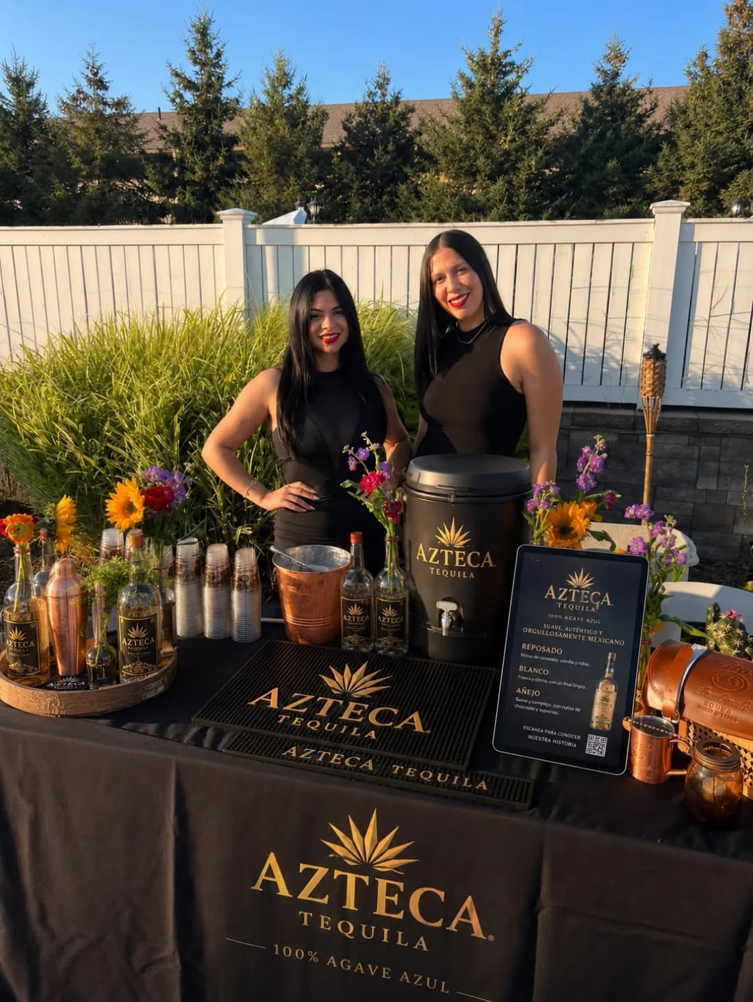 Two bilingual brand ambassadors at a tequila tasting activation — a branded table with bottle lineup, sampling kit and tasting tablet at an outdoor event.