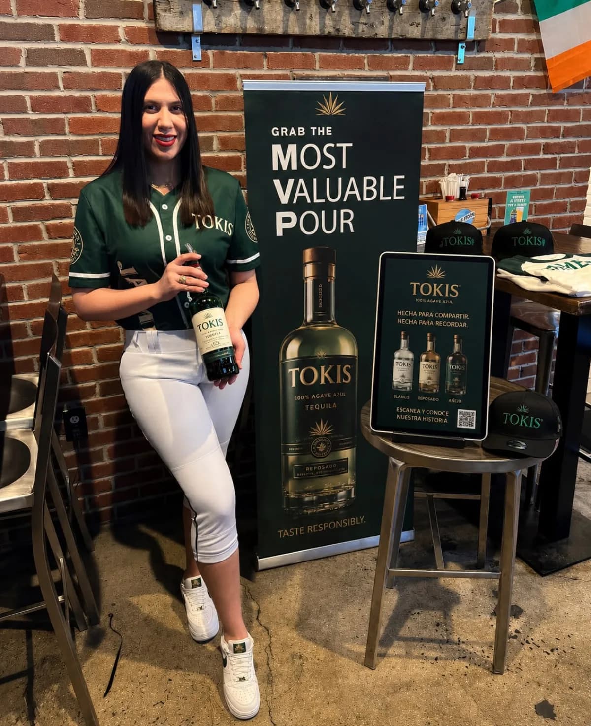 A bilingual brand ambassador at a tequila tasting activation, presenting the bottle beside a 'Most Valuable Pour' brand display and an interactive tablet.